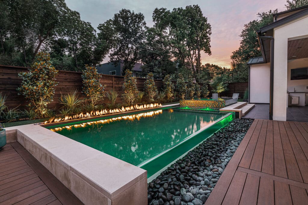 A modern backyard featuring a rectangular swimming pool with a water feature and built-in linear fire element along the far edge. Green lights illuminate the water. The area is surrounded by lush greenery, a wooden deck, and a pool house at sunset.