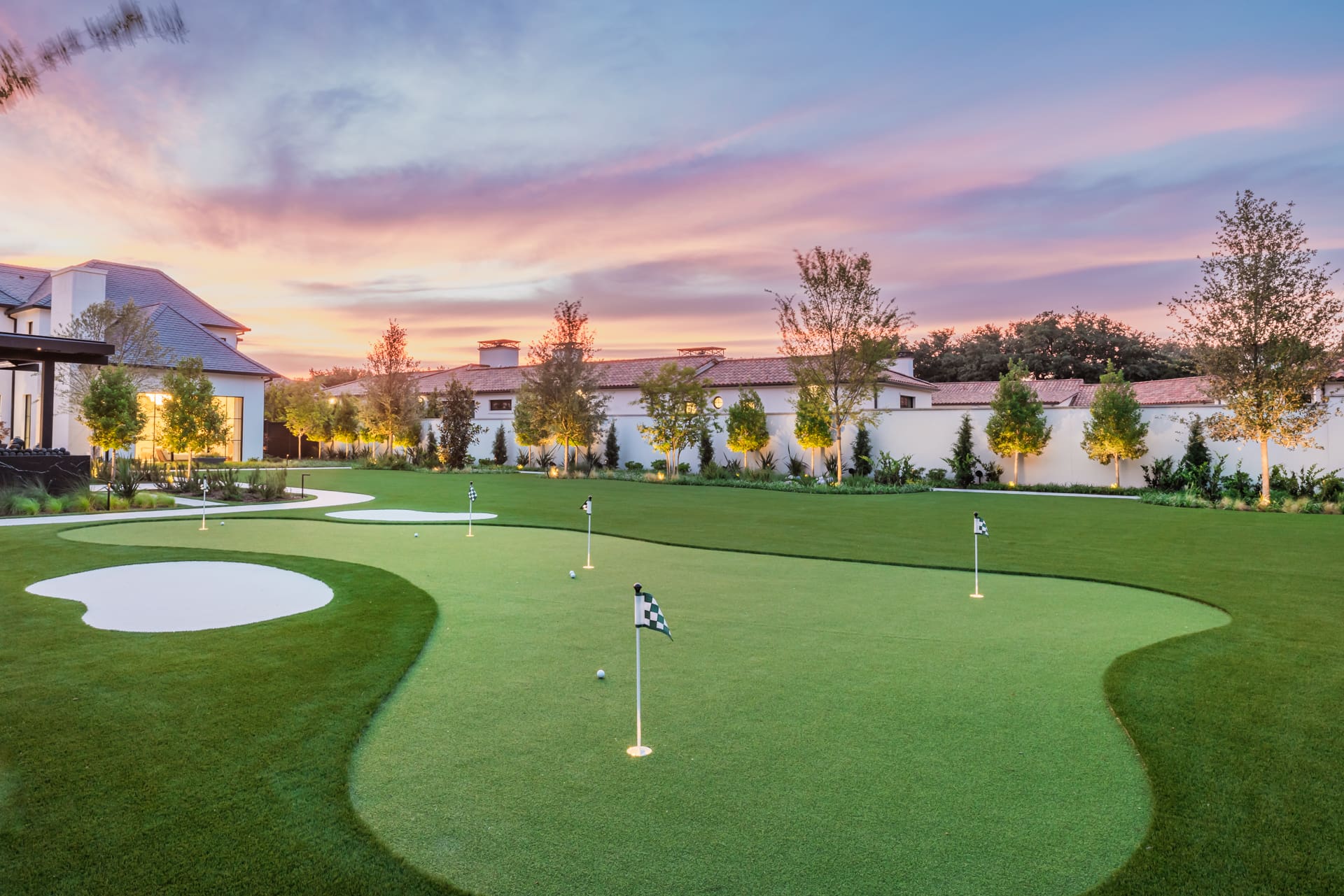 A beautifully landscaped golf putting green at sunset, with several holes marked by checkered flags. The lush green grass is surrounded by neatly trimmed trees and bushes. Modern buildings with red-tiled roofs are visible in the background under a colorful, cloudy sky.