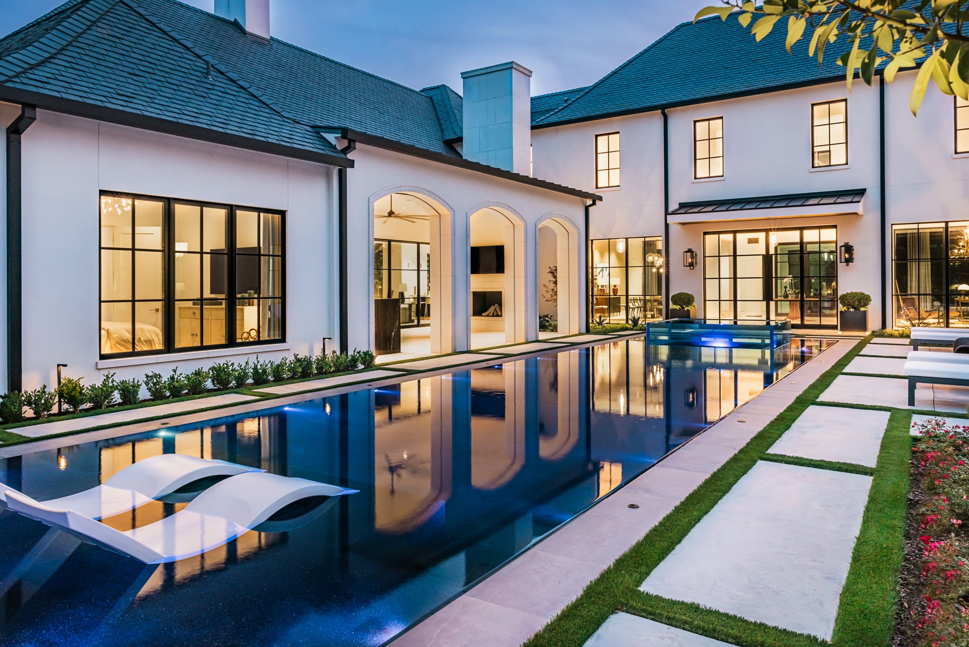 Luxurious modern house with large windows, a swimming pool with lounge chairs, and a well-maintained garden. The exterior features white walls and a dark roof, with well-lit interiors visible through the windows. Pathways and greenery surround the pool area.