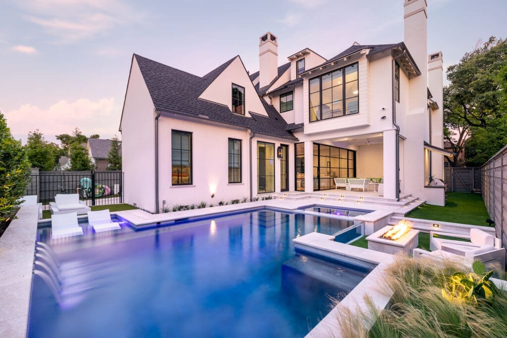 Modern white house with dark roof featuring a large, luxurious backyard pool surrounded by a patio. The pool area includes built-in seating, a hot tub, and a fire pit. Tall windows and greenery add to the elegant and tranquil atmosphere.