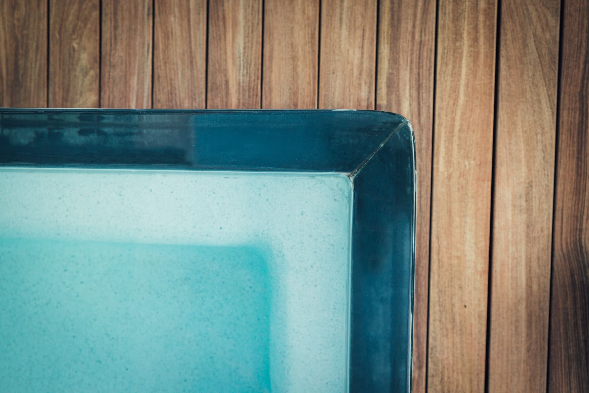 A close-up view of the corner of a glass-walled swimming pool filled with clear blue water. The backdrop features vertical wooden planks, adding a natural and warm contrast to the cool, clean lines and color of the pool.