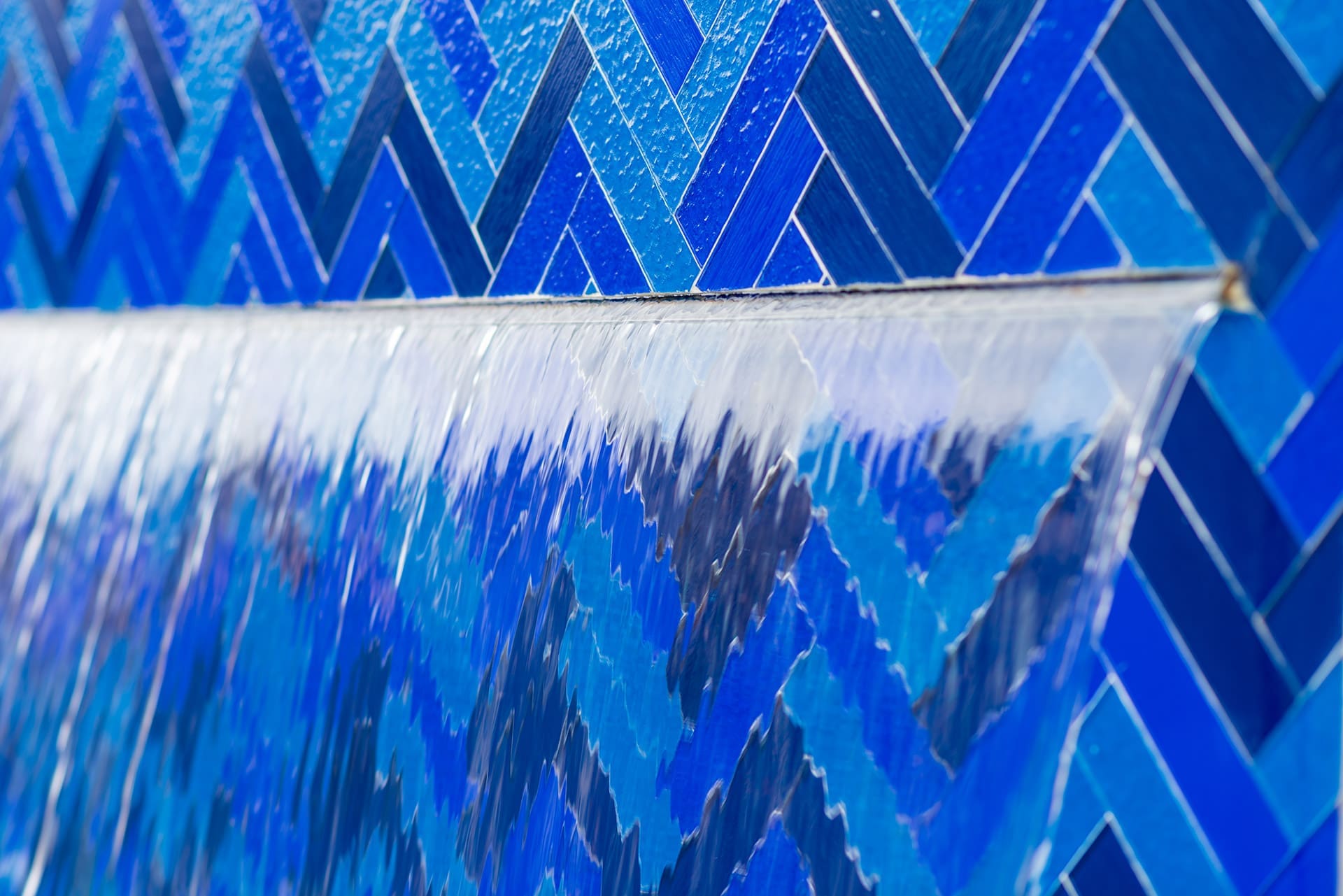 Water cascades over a smooth ledge, set against a background of blue, herringbone-patterned tiles, creating a reflective and visually captivating scene.