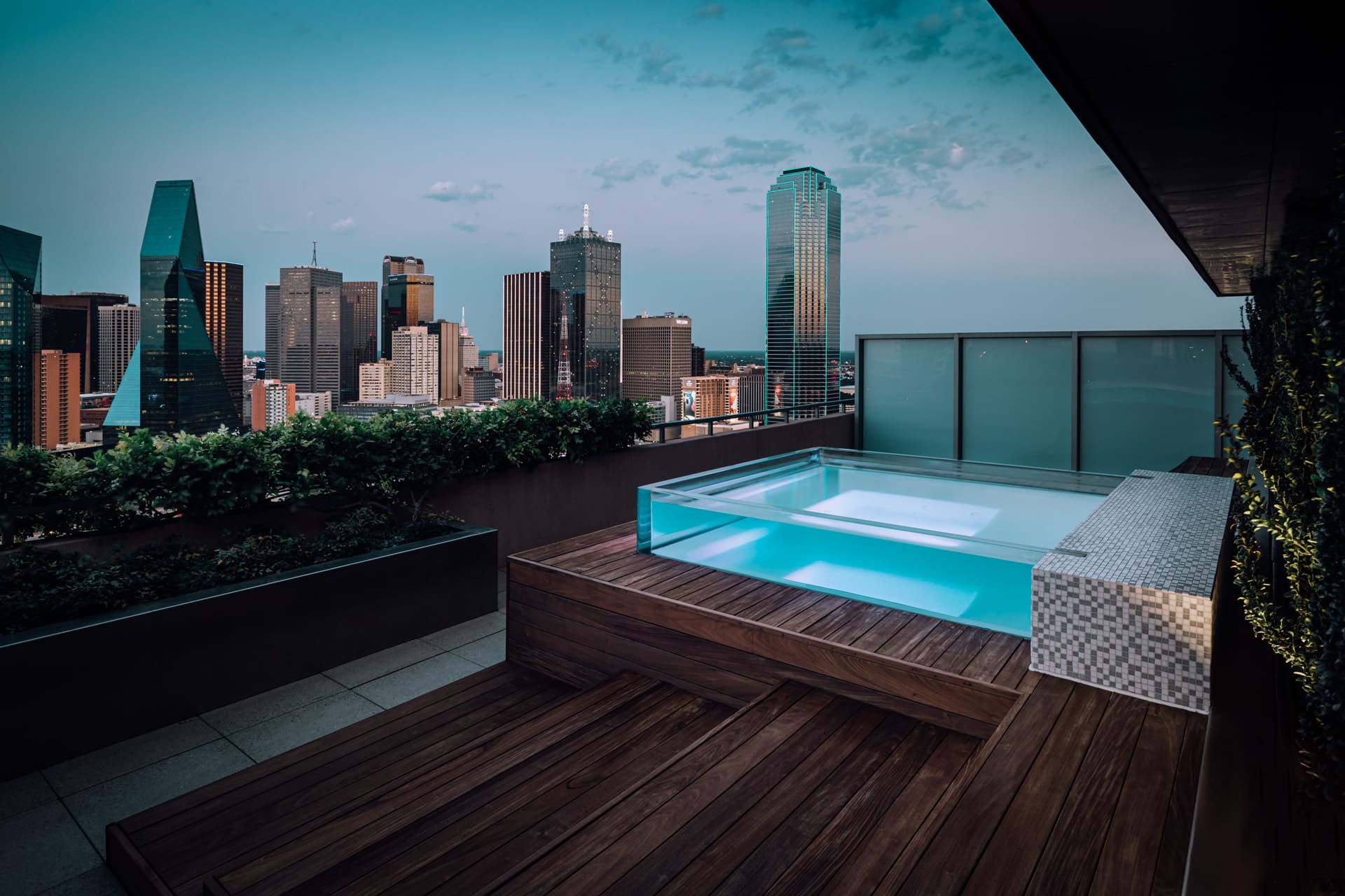 A rooftop patio features a modern wooden deck with an elevated, transparent edge, rectangular pool. Surrounding the space are lush green hedges and glass panels. The patio provides a panoramic view of a city skyline at dusk, with various skyscrapers in the background.
