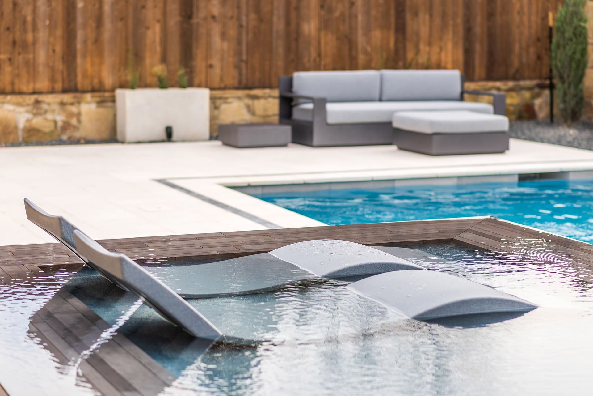 A modern pool area with sleek, gray lounge chairs partially submerged in shallow water is shown. In the background, there is a wooden fence, neatly arranged seating with gray cushions, and potted plants, creating a stylish and relaxing outdoor space.