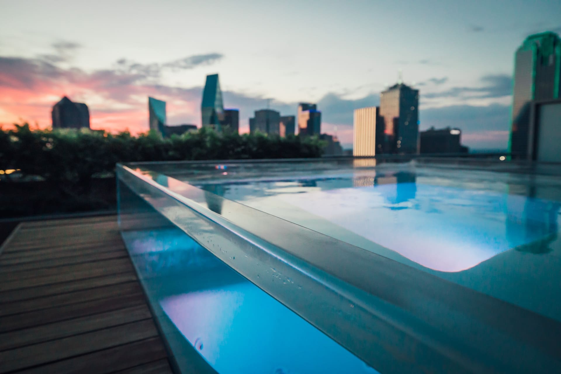 A rooftop pool with glowing blue lights overlooks a city skyline at dawn or dusk. The glass barrier around the pool reflects the serene water, while the city buildings in the background are silhouetted against the colorful sky.