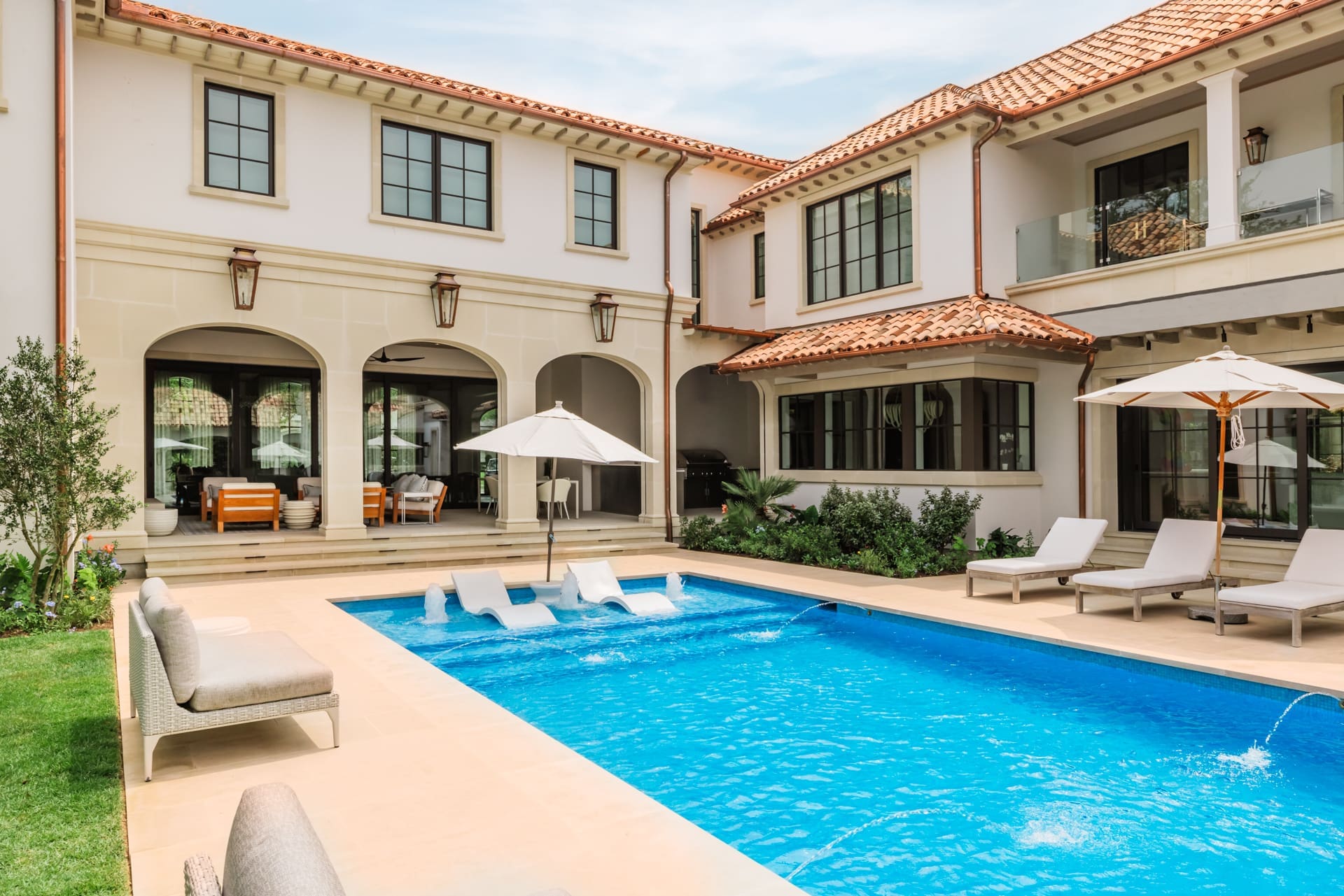 A luxurious Mediterranean-style villa with a terracotta roof features an outdoor patio, dining area, and a swimming pool with lounge chairs and umbrellas. The creamy stucco facade and large windows enhance the elegant atmosphere.