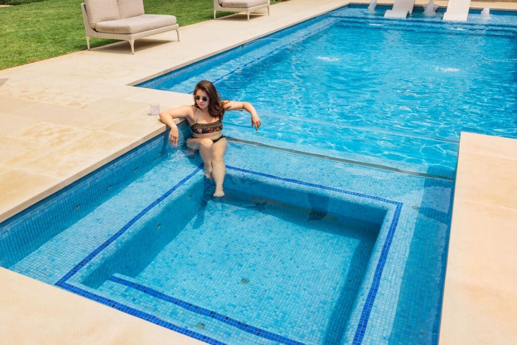 A woman in a black and orange bikini is lounging on the edge of a clear blue pool, resting her arm on the ledge and her feet in the water. She is wearing sunglasses, and there are lounge chairs and grass nearby.