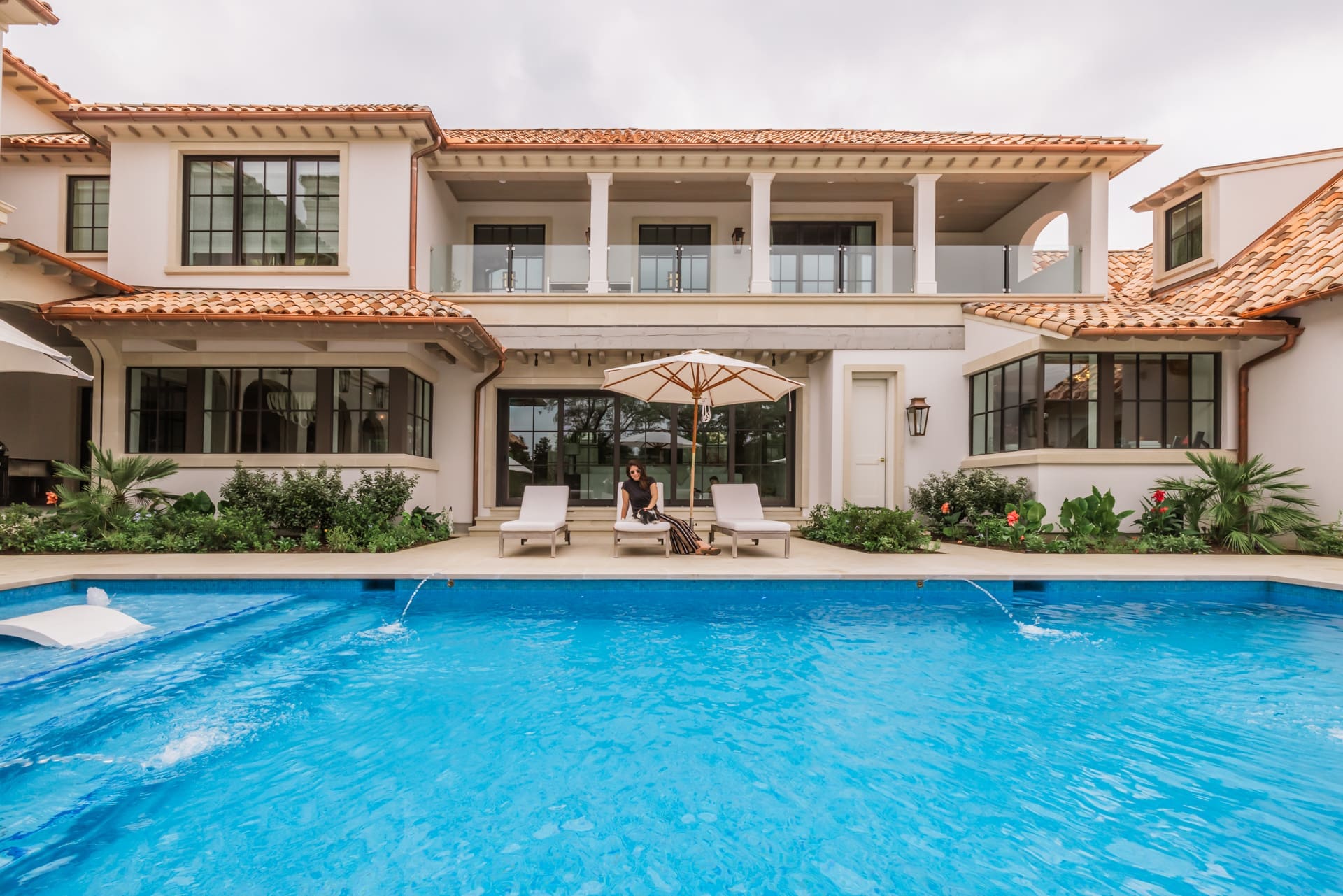A luxurious two-story house with a red-tiled roof is seen from the backyard. It has large windows and a spacious balcony. In front, there's a swimming pool with two white lounge chairs and a large umbrella beside it. Lush greenery surrounds the area.