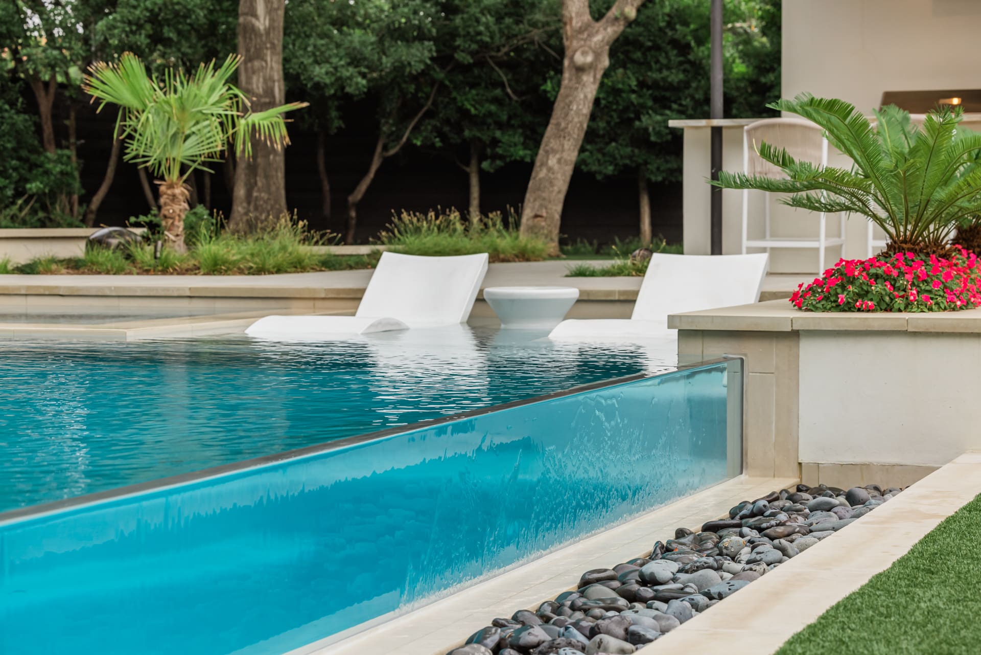 A modern pool with a transparent edge, surrounded by smooth stones. Two white loungers and a small table sit partially submerged. There are lush plants, a row of trees, and a small white bar area in the background.