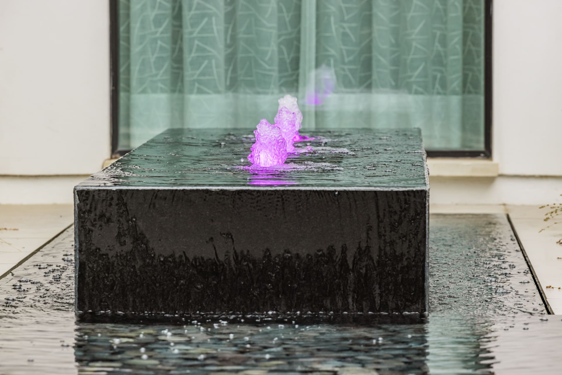A modern rectangular black stone fountain with glowing purple water jets is set against a backdrop of tall glass windows with patterned curtains. The fountain is surrounded by a shallow pool of water.