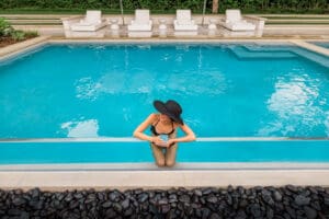 A woman in a black swimsuit and wide-brimmed hat stands in a swimming pool, holding a drink at the pool's edge. White lounge chairs line the poolside in the background.