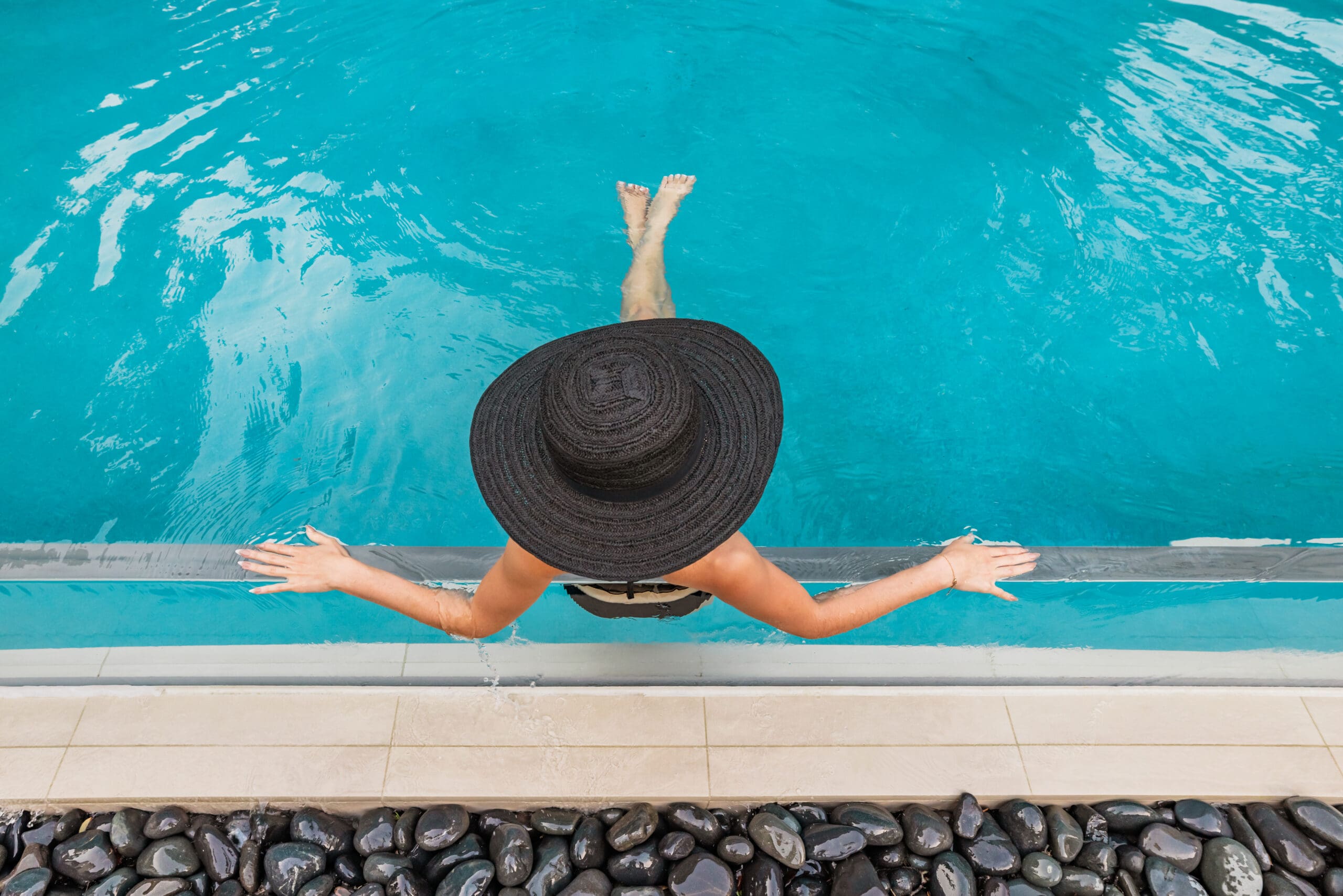 A person wearing a large black sunhat relaxes at the edge of a swimming pool, sitting on the side with their feet in the water and hands resting on the poolside. The image is taken from above.