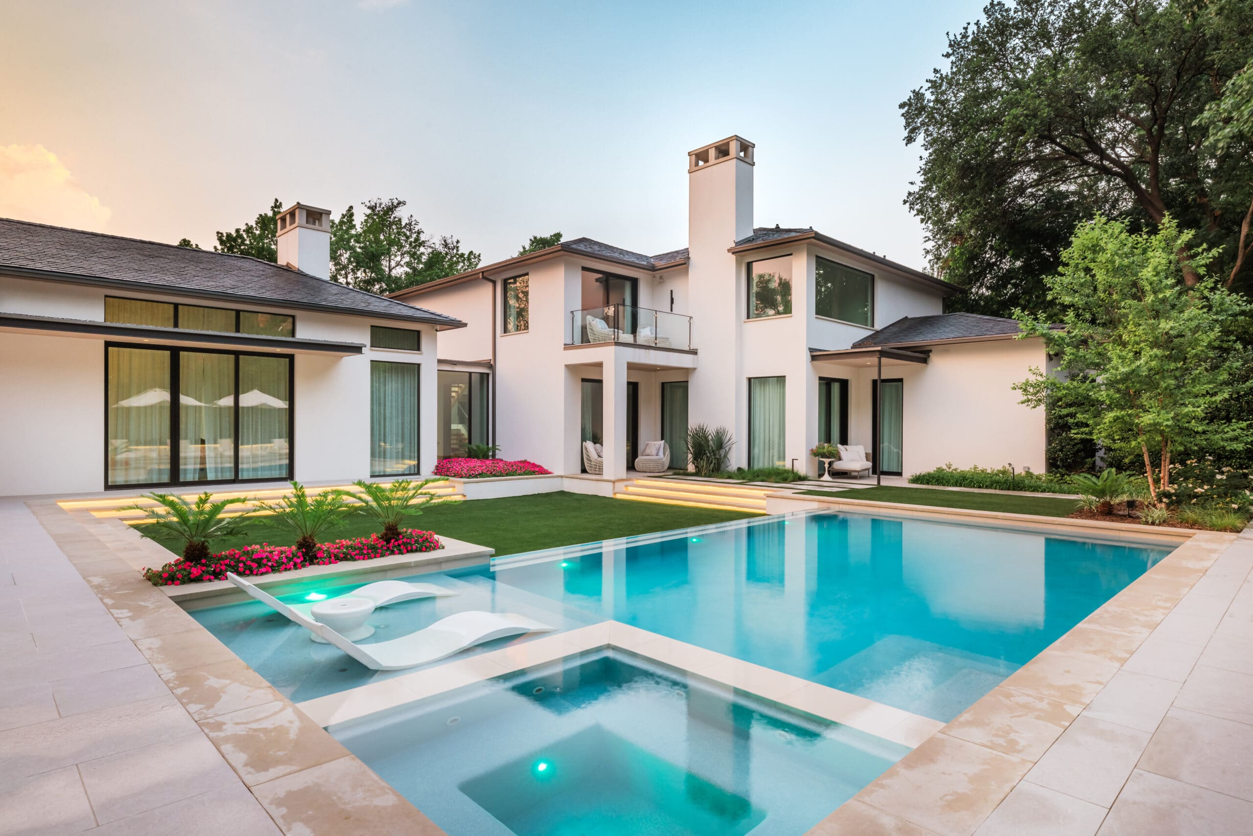 Modern two-story house with large windows, surrounded by trees, featuring a luxurious backyard with a swimming pool, spa, sun loungers, green lawn, and vibrant flower beds.