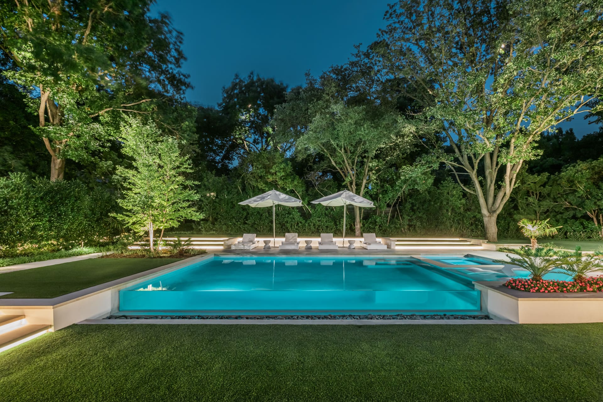 A serene outdoor pool area at dusk, surrounded by trees. The illuminated pool reflects soft lighting, with sun loungers and umbrellas in the background. A manicured lawn and flower bed enhance the peaceful ambiance.