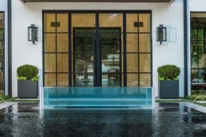 Modern glass-walled pool with clear water, surrounded by a black-tiled deck. Behind the pool are large grid-style glass doors, potted plants, and a white building with wall-mounted lanterns.