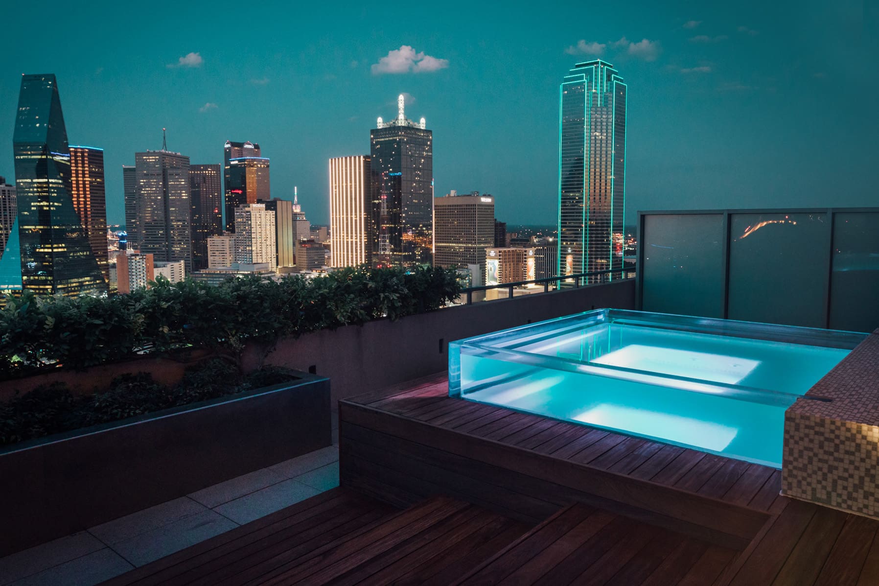A rooftop pool with clear walls glows blue at night, overlooking a city skyline with tall, illuminated buildings under a dusky sky.