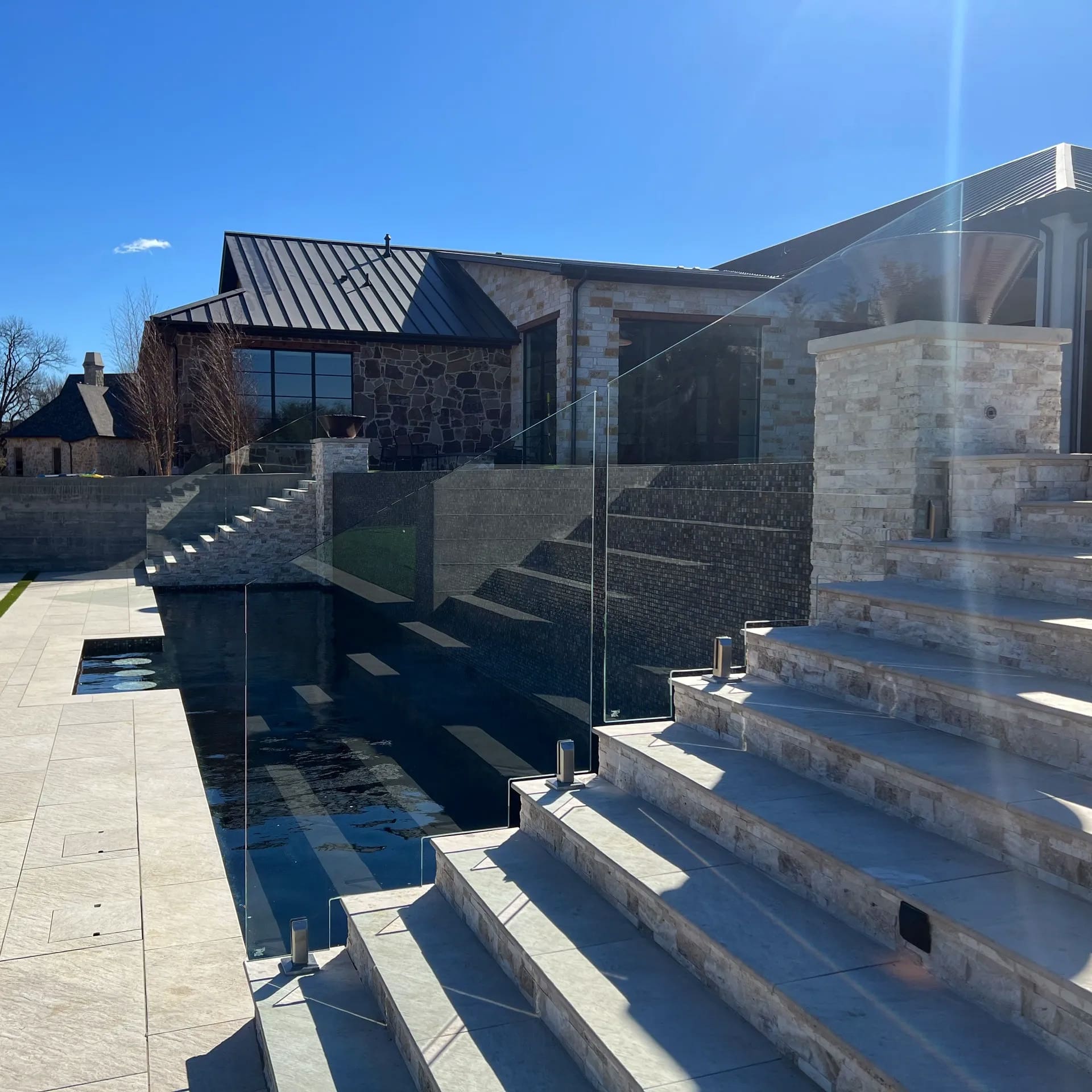 Modern outdoor area with wide stone steps, glass railing, and a sunken pool leading to a contemporary house with dark metal roof and stone exterior under a clear blue sky.