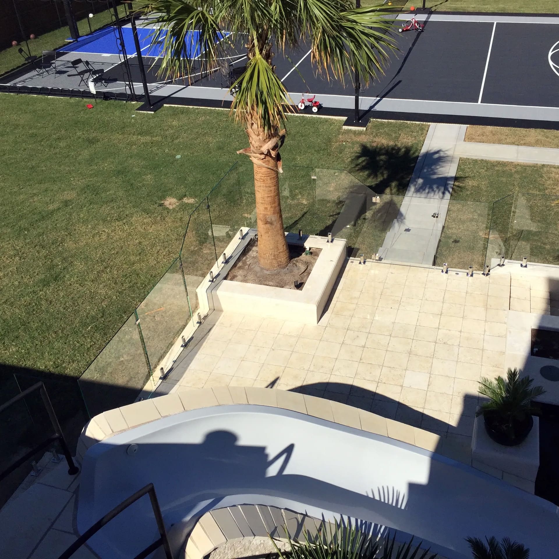 A backyard view featuring a tiled patio with a palm tree, a curved white slide, and a fenced sports court with tennis or pickleball markings in the background. Grass surrounds the patio and court.
