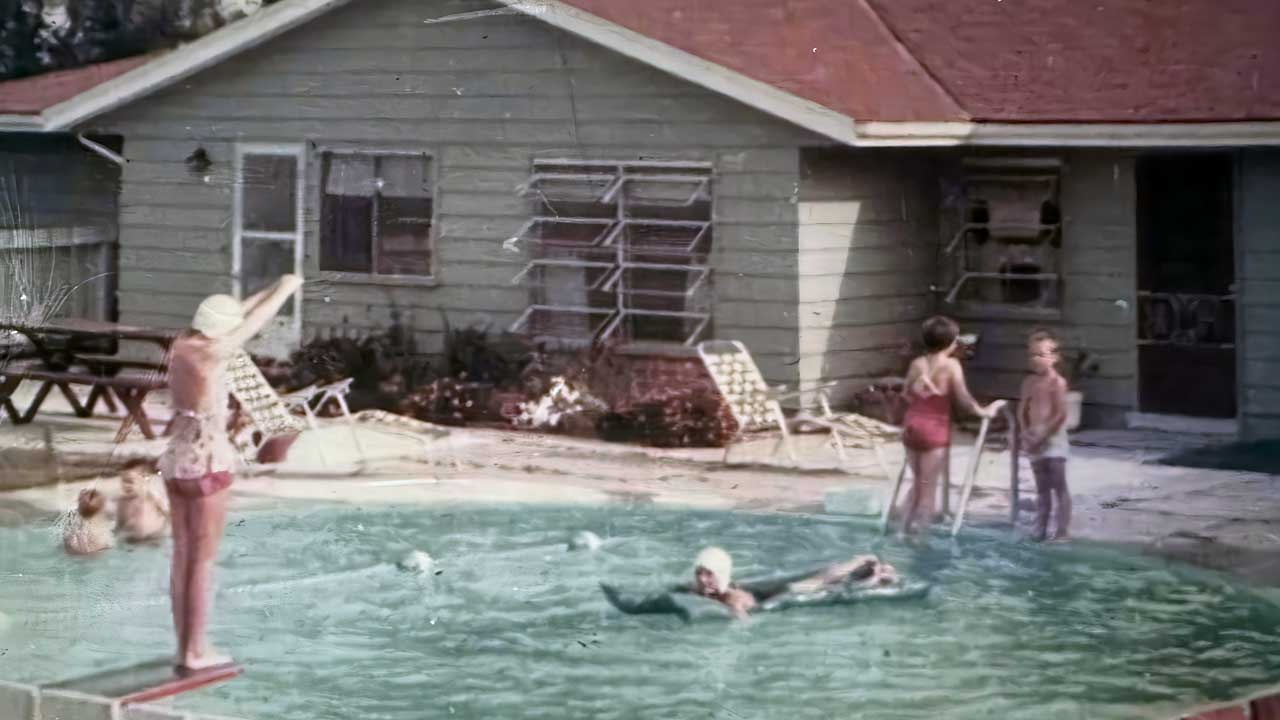 Several children play in a backyard pool near a light green house with a red roof. One child stands on a diving board, and others swim or stand near the pool ladder. Lounge chairs and flowers are visible by the house.