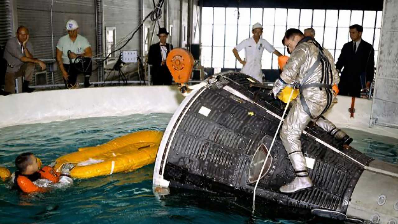 An astronaut in a silver suit stands on a space capsule floating in a pool, while another person in an orange suit floats nearby with an inflated life raft; several people watch from the side.