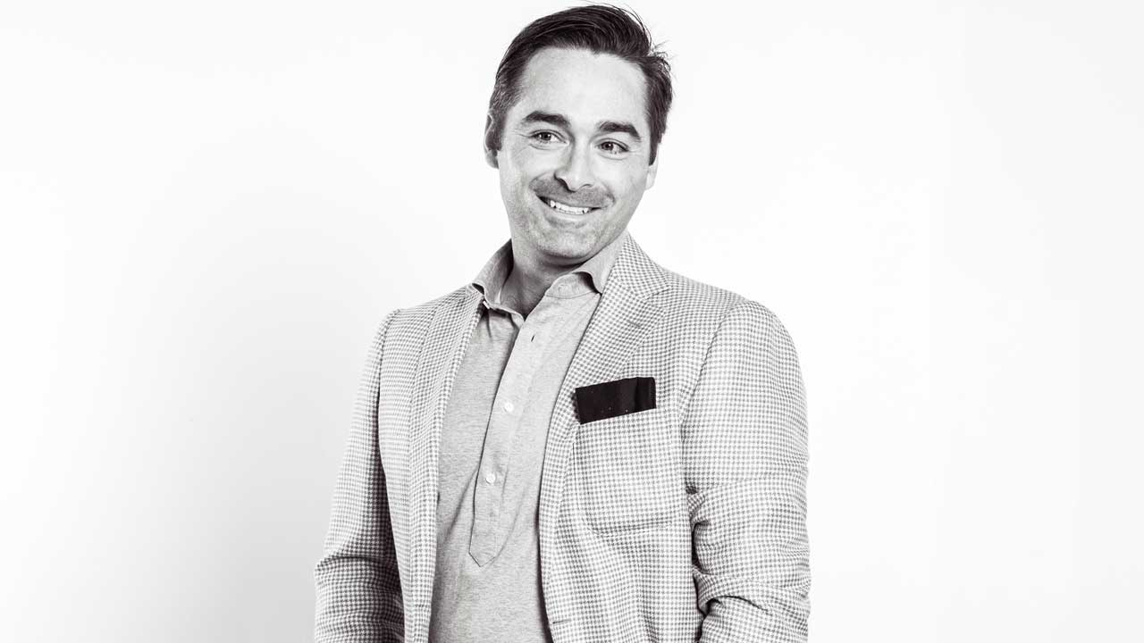 A man in a light-colored checkered blazer and polo shirt smiles while standing against a plain white background. A pocket square is visible in his blazer pocket.